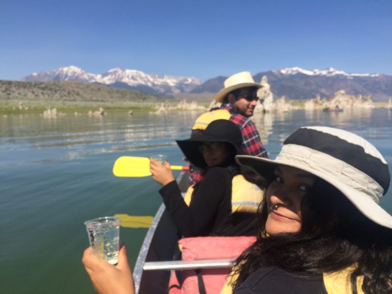 Three students sitting in a canoe on Mono Lake looking at the camera with clear plastic cups filled with Mono Lake water in their hands, smiling on a beautiful day with glassy green and blue water around them and tufa towers and mountains in the distance.