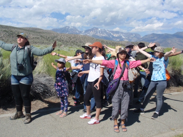 A tour guide stands in front of a group of students on a trail outside on a beautiful sunny day and they are all facing the same direction with their arms outstretched as if hugging the world.