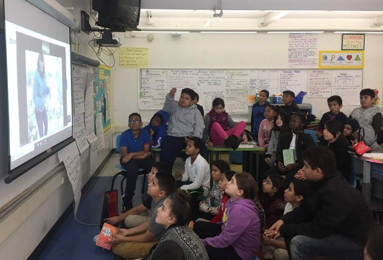 A classroom with 23 students looking intently at a large projection screen showing a person giving a live tour at Mono Lake.