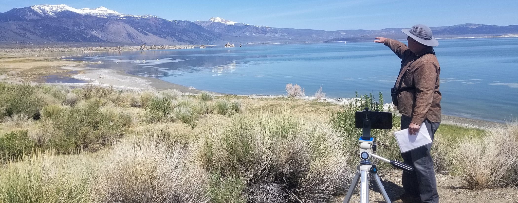 A man is outside with a hat and binoculars pointing out something in the distance while he stands in front of a cell phone mounted on a tripod so he is giving a virtual tour overlooking Mono Lake.