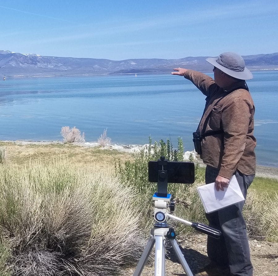 A man is outside with a hat and binoculars pointing out something in the distance while he stands in front of a cell phone mounted on a tripod so he is giving a virtual tour overlooking Mono Lake.