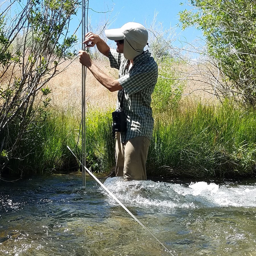 Field technician stands knee deep in a stream with a tall instrument that he is holding vertically in the stream with a long measuring tape going from across the stream bank, and there are green grasses and willows and rushing water.