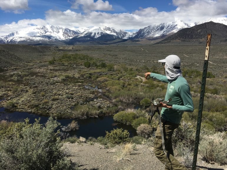 A person stands on a bluff overlooking a wide stream system with water flowing and green vegetation below and snow-capped mountains in the distance and he has a wildlife camera in his hand and is pointing to a place off in the distance.