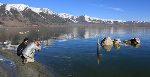 Two people kneel at the shore of Mono Lake and look out, one with binoculars, at a lake level gauge just off shore, with a glassy blue lake and snow-capped mountains in the background.