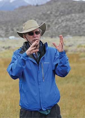 A man, Peter Vorster, in a sun hat and sunglasses stands in a field gesticulating with his hands while speaking.