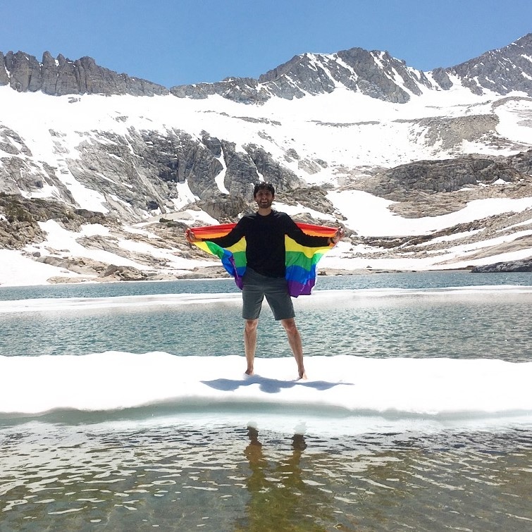 Andrew Youssef standing on a floating piece of ice on a mountain lake with a rainbow Pride flag in his outstreached arms.