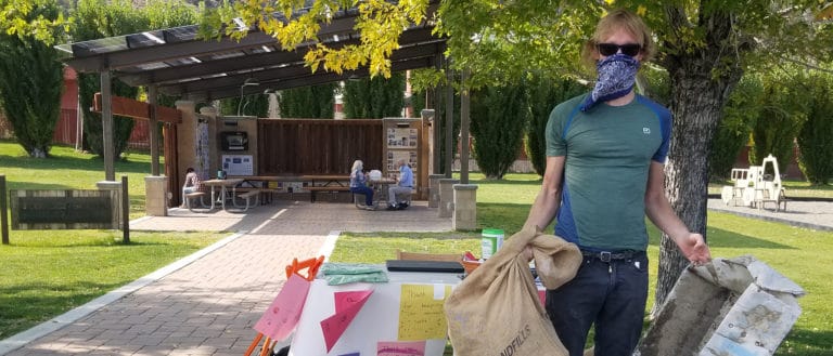 A person wearing a face mask holds a trash bag and some recycling in front of a table set up for a trash-collecting event.