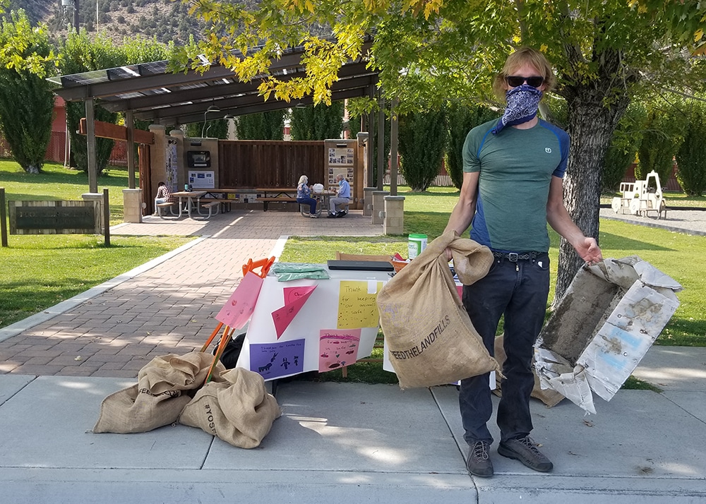 A person wearing a face mask holds a trash bag and some recycling in front of a table set up for a trash-collecting event.