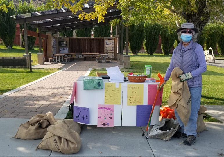 A woman in a face mask and gloves poses in front of a table with a trash collecting tool and a canvas bag.