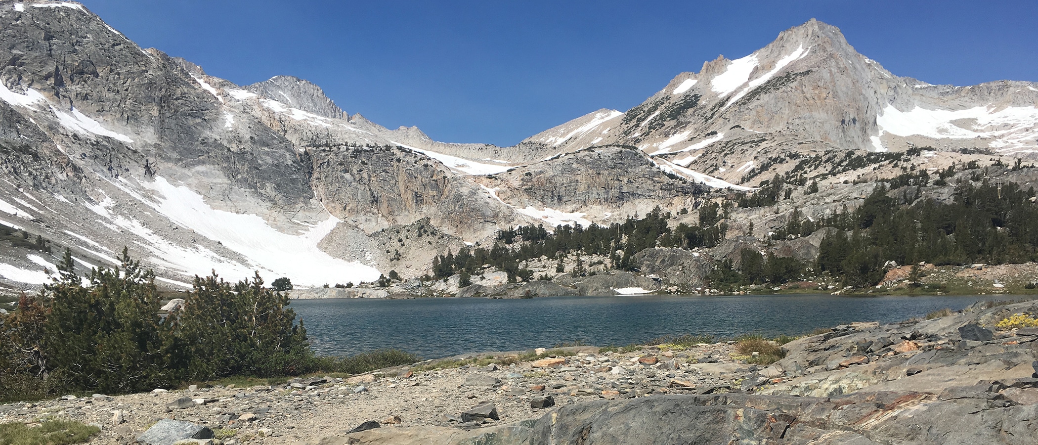 Blue Saddlebag Lake in the foreground with high alpine granite mountains with patches of snow in the background.