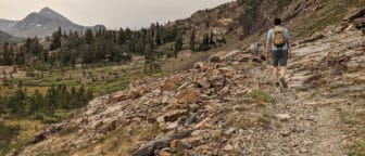 Hiker with a backpack walks along a rocky trail up in the mountains with alpine trees and a sharp Sierra skyline.