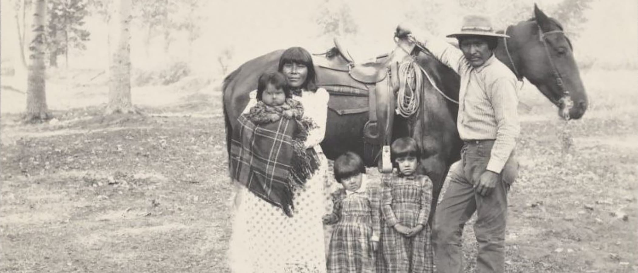 A Kutzadika'a family with a Mother, baby in arms, two young firls, and a Father stand in front of a saddled horse.