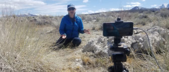 A person in a blue jacket sits in the dry grass and talks to an Iphone which is propped up on a tripod.