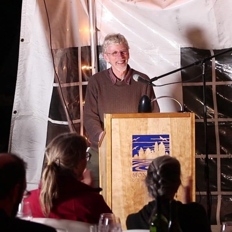 A man, Peter Vorster, stands at a lecturn, smiling, inside of an event tent with people looking on.