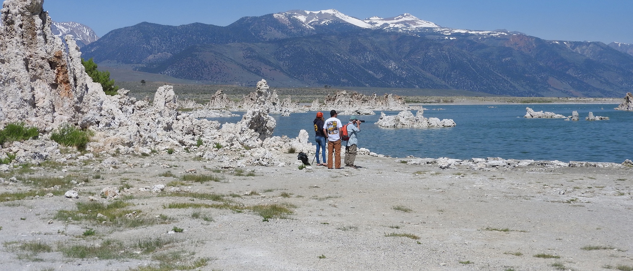 Three people walking along the shore of Mono Lake with tufa towers around them and in the water, with the snowy Sierra Nevada mountains in the background.