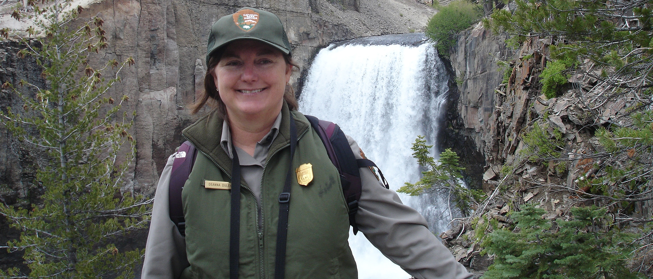 Woman in a National Park Service vest and ball cap standing in front of a large waterfall.