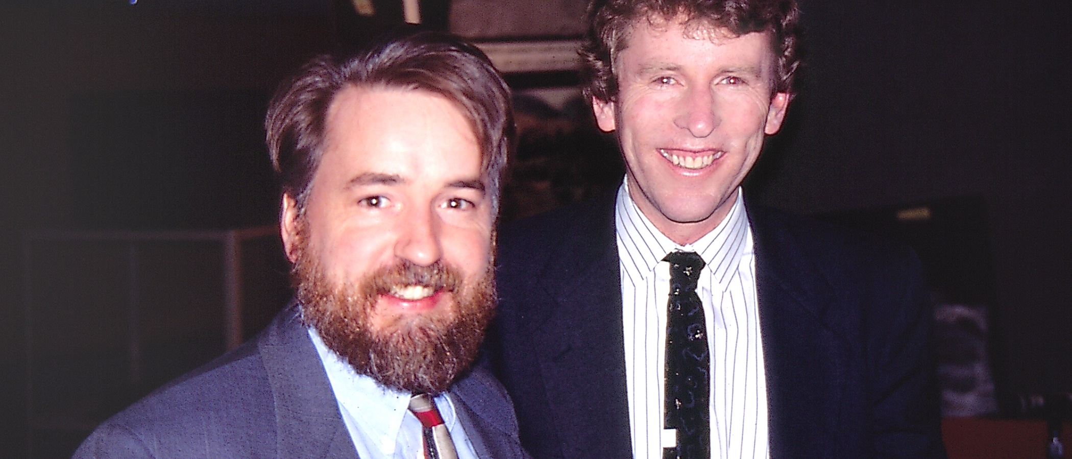 Two smiling people standing next to each other wearing suits and ties.