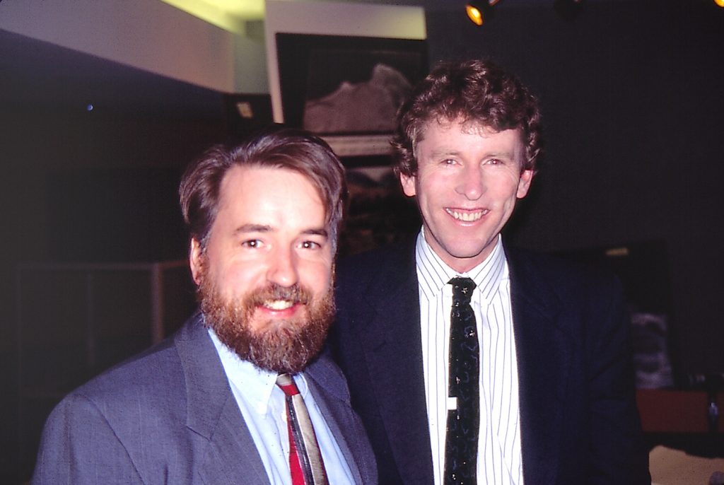 Two smiling people standing next to each other wearing suits and ties.