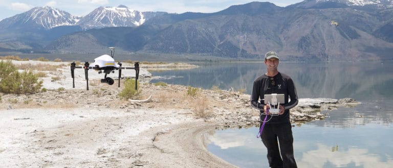 A research drone hangs in the air next to a person holding the drone controls on a bright sunny day at Mono Lake with the lake and mountains in the background.