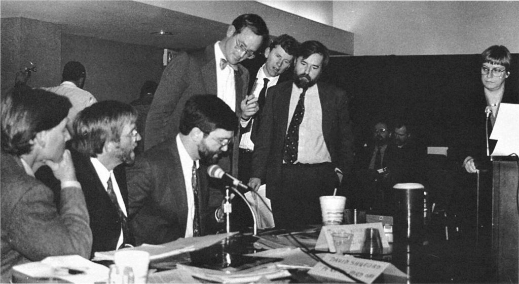 Black and white, a group of men in suits, three sitting and three standing, deliberate at a table strewn with papers. A woman standing at a podium to the right looks at them.