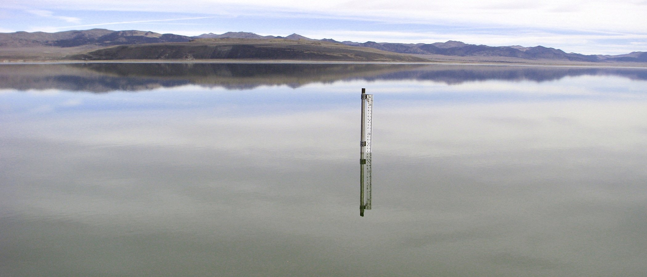 A lake level gauge, made of a metal pipe with a ruler-like measuring stick attached, pokes up from the surface of a very glassy and highly reflective lake with the lenticular-shaped Black Point and desert mountains in the background.