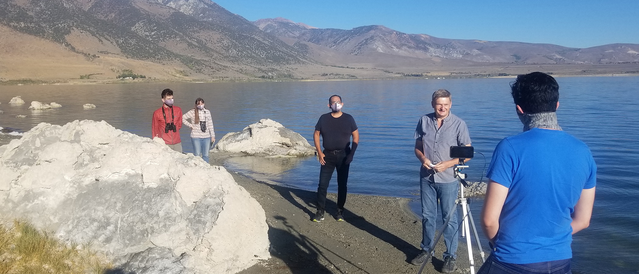 Five people standing at the shoreline of Mono Lake, socially distanced and masked, shooting a video from a cell phone on a tripod with a glassy Mono Lake and the Sierra Nevada behind them.