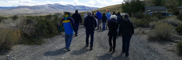 A group of people walk away from the camera down a gravel road through light green and darker green sagebrush. The Sierra Nevadas sit in a cloudy sky.