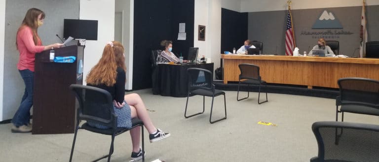 Woman stands at a podium in a hearing room with socially distanced chairs and people sitting at computers with the United States and California flags.