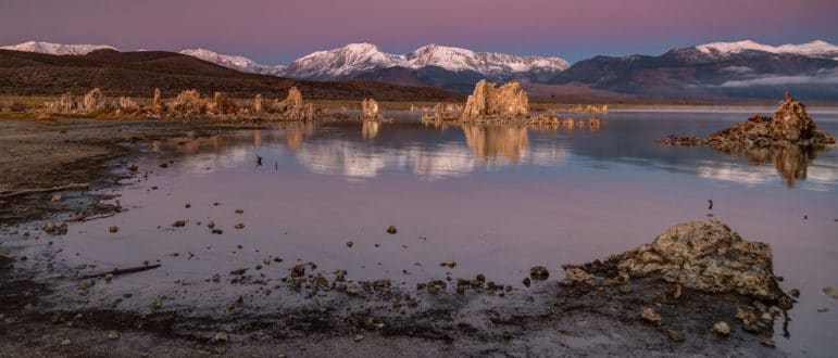 Shoreline of Mono Lake looking west towards the Sierra Nevada with tufa towers reflecting on the glassy lake and the sky is pink, purple, and blue.