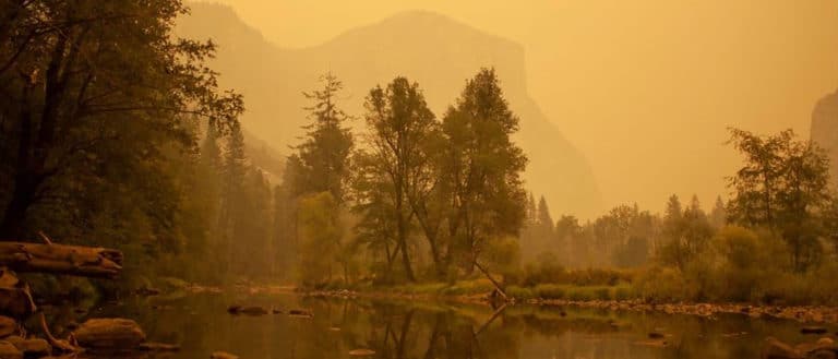 View of the Merced River flowing through a very smokey Yosemite Valley with orange smoke hanging in the air and coloring everything in muted hues.