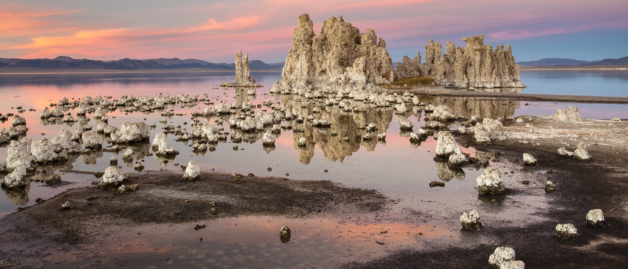 A pink and blue sunset fills the sky and is reflected in the glassy waters of Mono Lake. Small pieces of tufa poke out of the water and eventually lead to a group of tufa towers in the center.