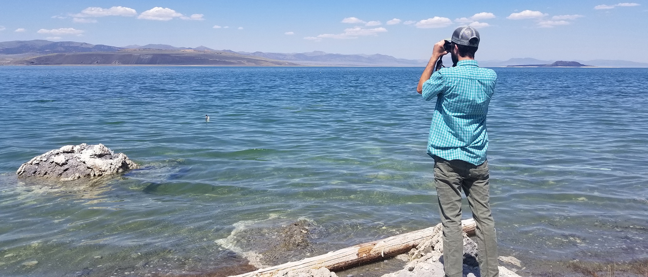 A man in a blue shirt faces away from the camera on the edge of clear and wavy turquoise water, and looks through binoculars.