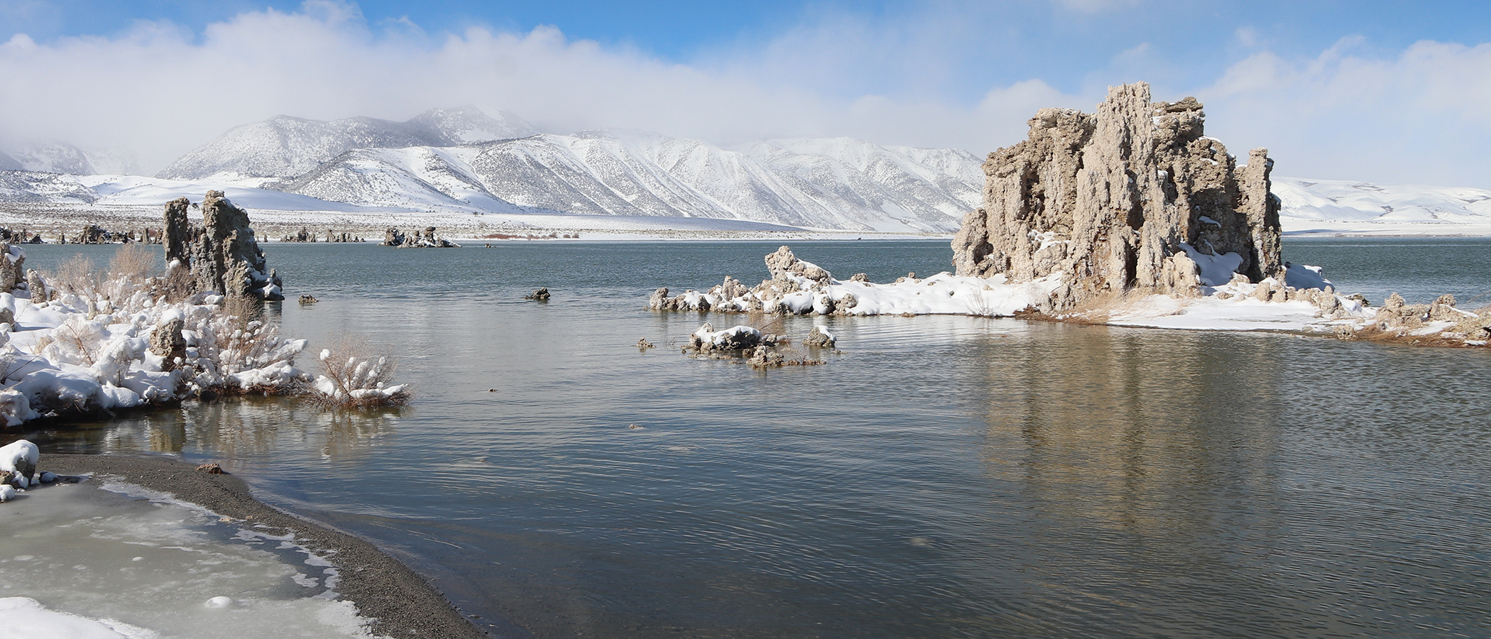 Majestic light brown tufa towers jut out of dark water with snow covered mountains and a blue sky behind them.