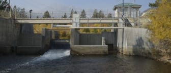 Water flows through several concrete structures, which connect to a gray metal railing and stairway, as well as a light yellow bridge over the creek. Yellow foliage lines the water.