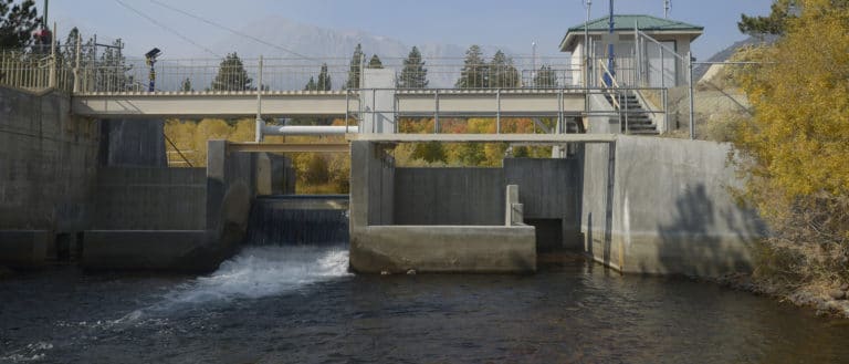 Water flows through several concrete structures, which connect to a gray metal railing and stairway, as well as a light yellow bridge over the creek. Yellow foliage lines the water.