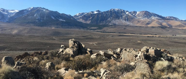 A brown hilly landscape dotted with evergreen trees slopes up into the dark and snow striped Sierra Nevada mountains against a blue sky.