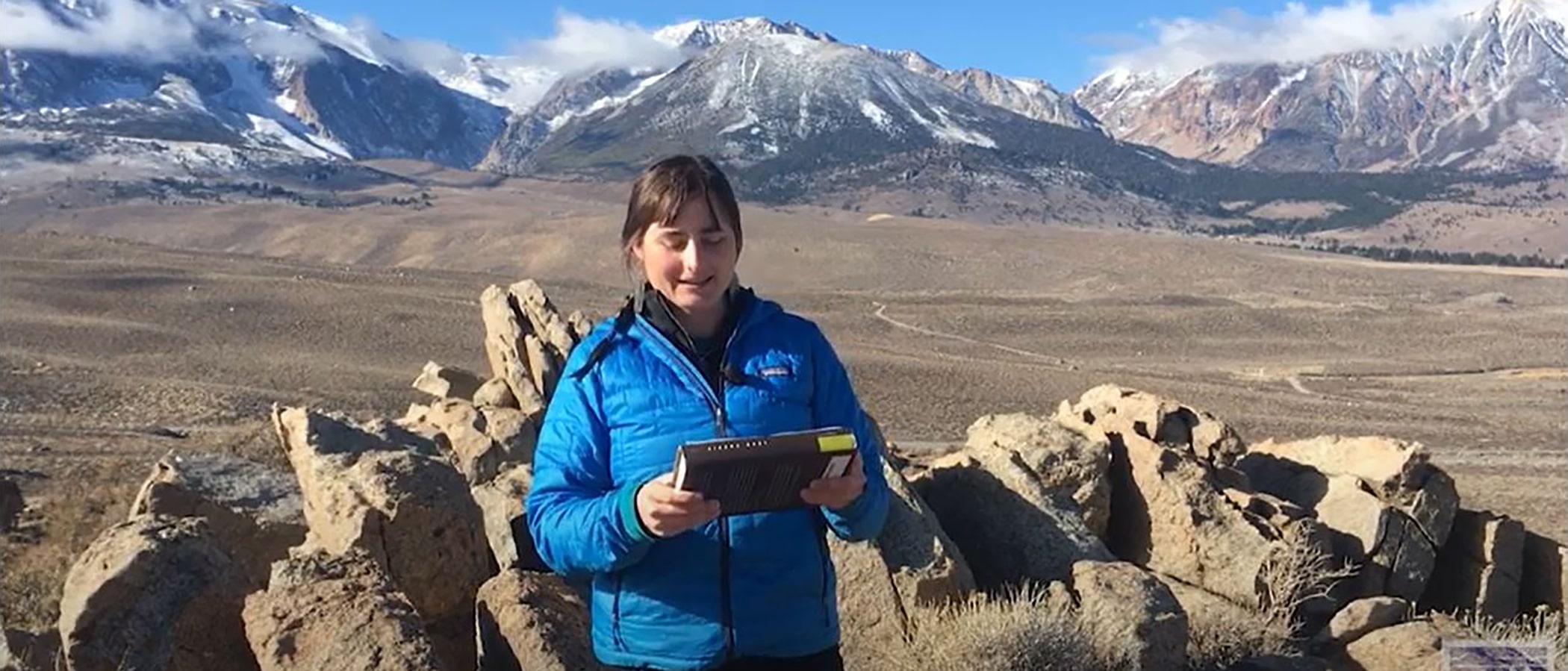 A woman with brown hair and a blue coat stands in front of a rocky vista looking out on brown fields and mountains. She is holding a book.