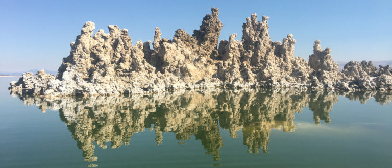 A large group of tall, light brown tufa towers juts our of the dark green water into a cloudless blue sky. The towers' reflection ripples below.