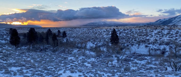 a snow speckled valley stretches ahead, bordered by mountain's edges, and the glowing orange and pink sunset peeks out behind long fluffy clouds.