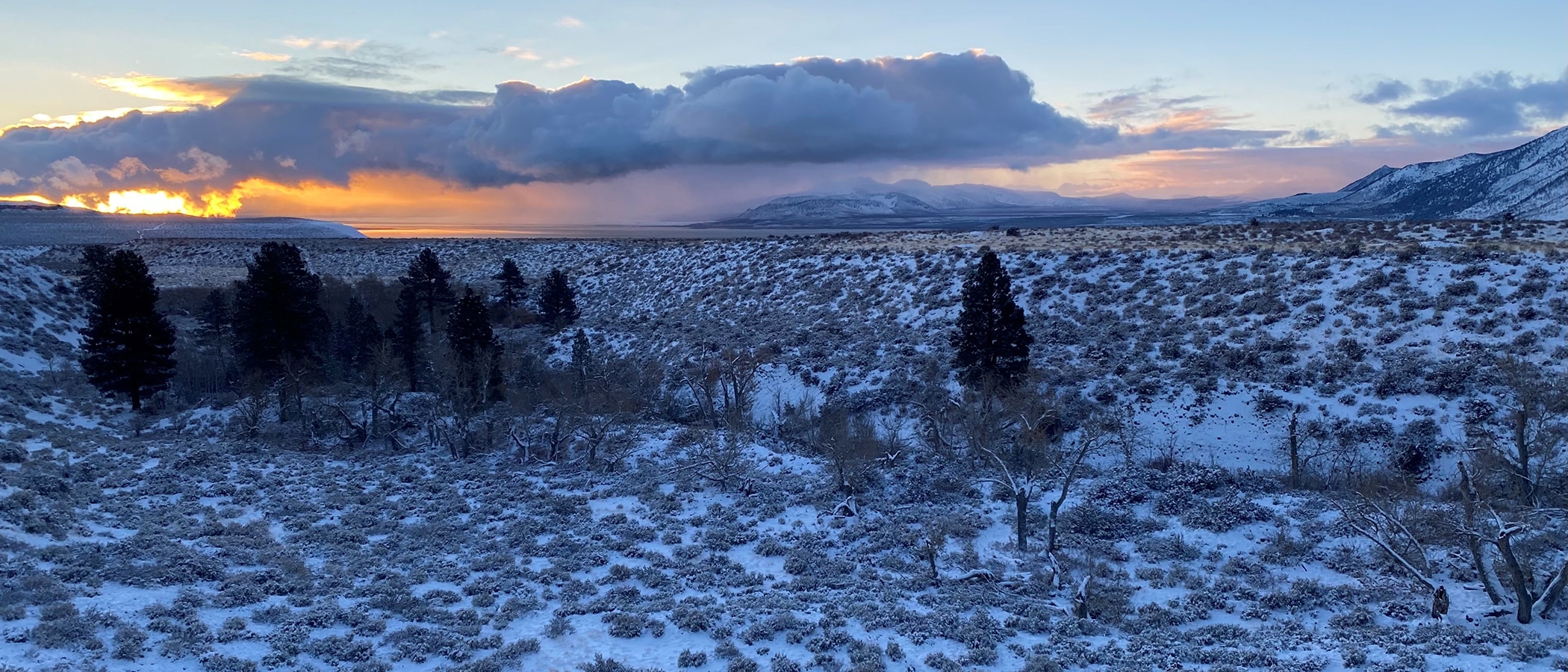 a snow speckled valley stretches ahead, bordered by mountain's edges, and the glowing orange and pink sunset peeks out behind long fluffy clouds.