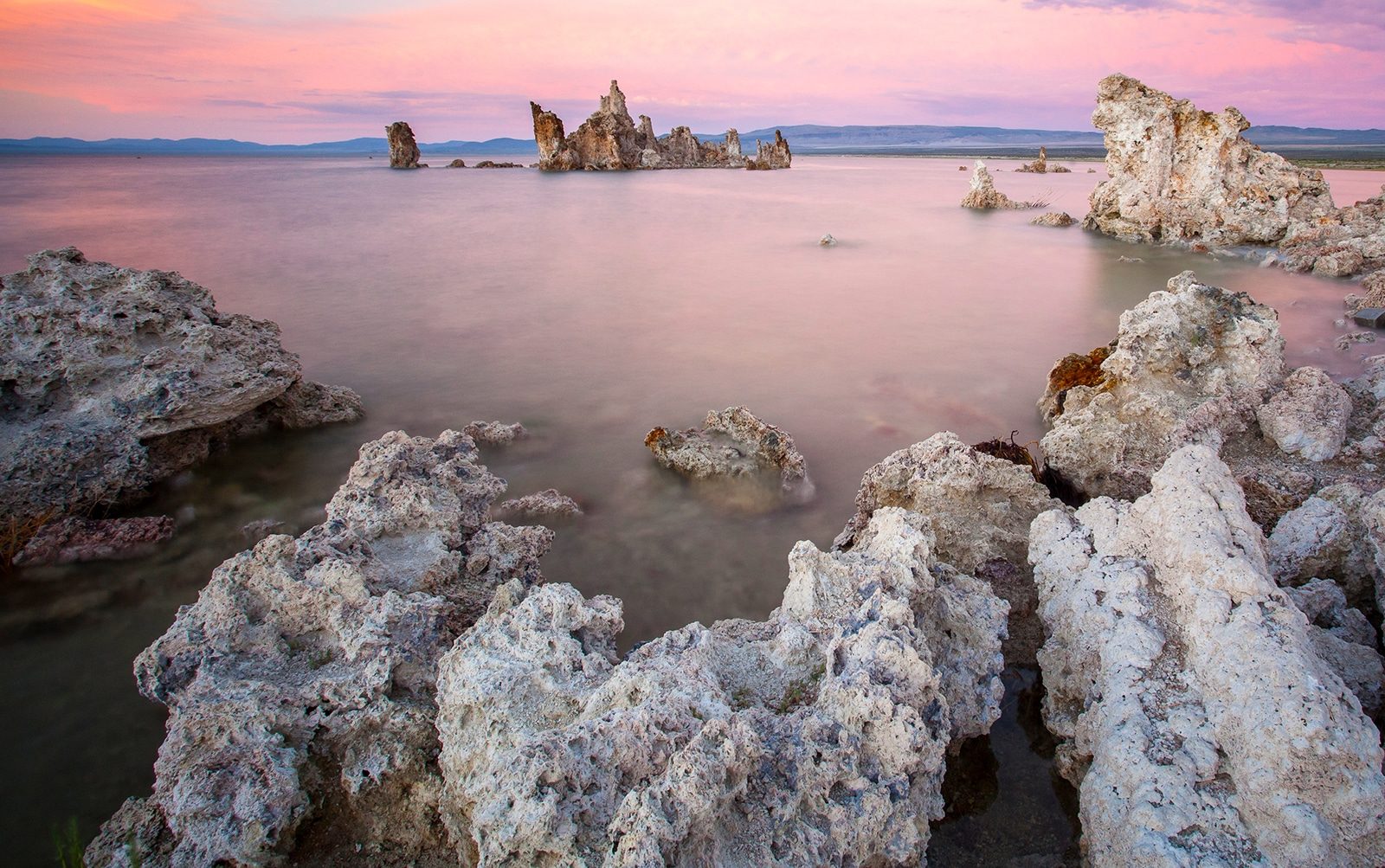 Sunset at Mono Lake with pink hues in the clouds and reflected in the water. with an island made of tufa in the distance and tufa towers framing the foreground.