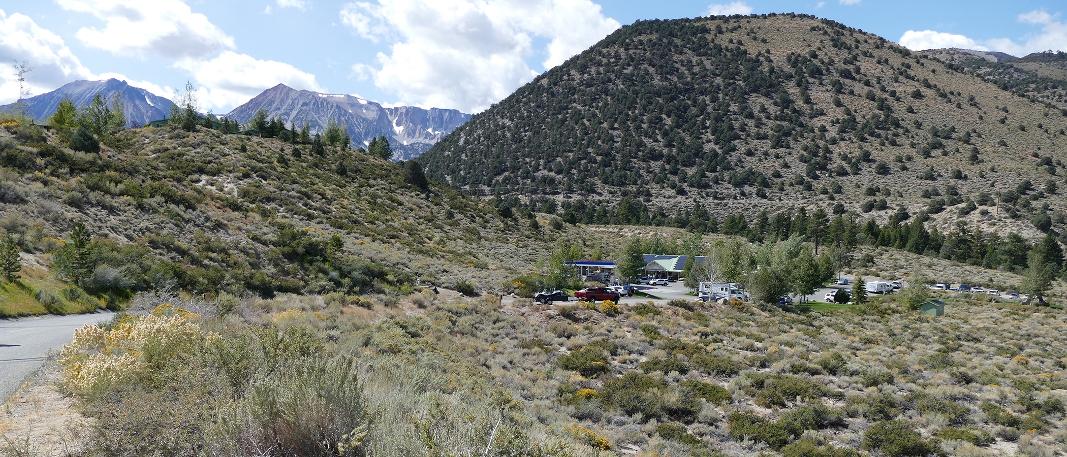 Trees and shrubbery colored light and dark green line the hills in the foreground, and rocky dark blue and snowcapped mountains are peaking out behind them.