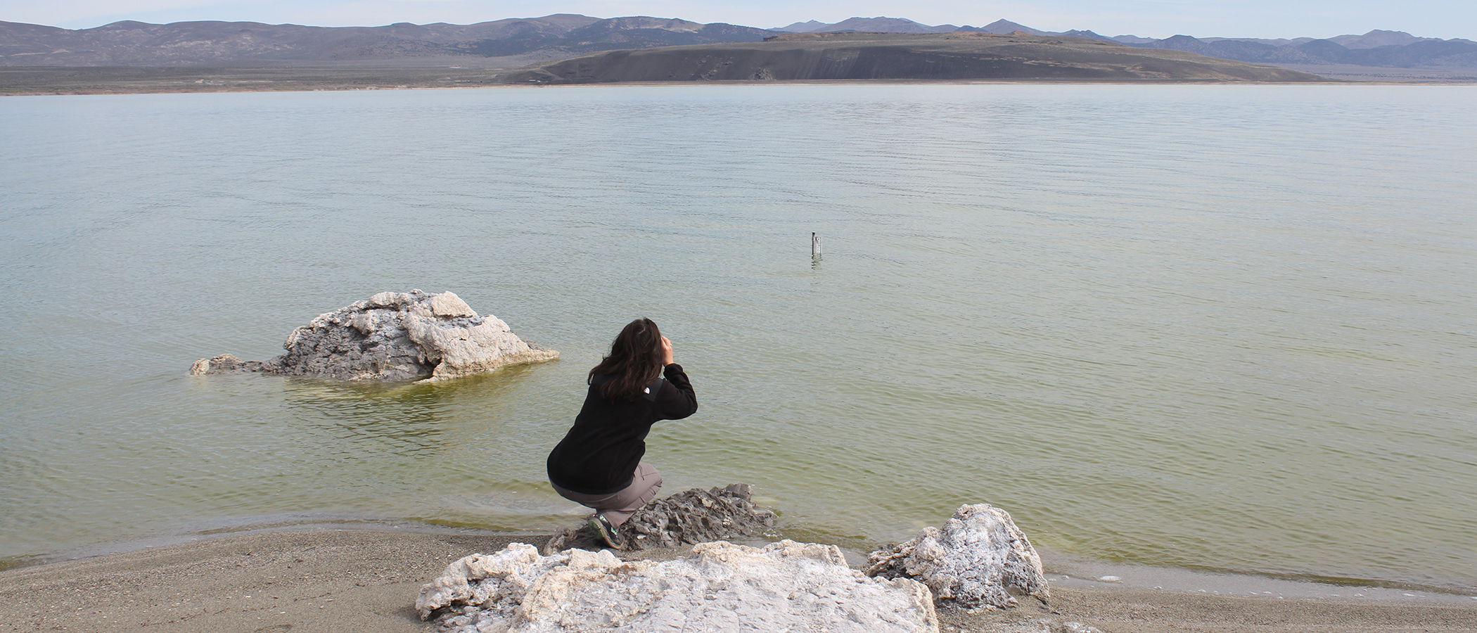 A woman crouches at the gray blue edge of Mono Lake and looks out towards the large black rock outcropping on the opposite shore.