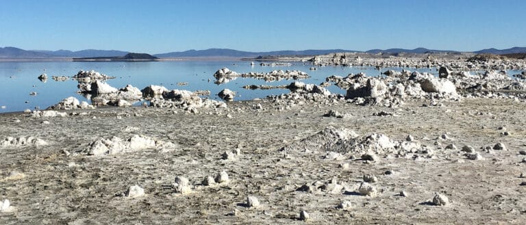 Small rocky tufa pieces are scattered across a beach and in the blue water, which reflects a clear sky.