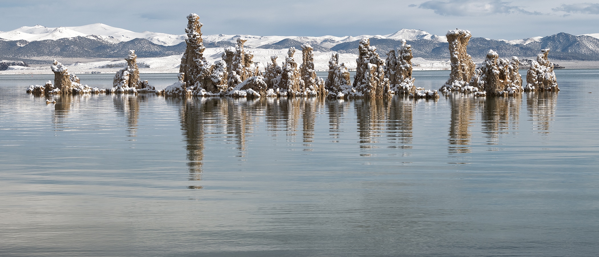 Snowy tufa towers jut out from the rippling green lake water with the snowy Sierra Nevada mountain range behind them.