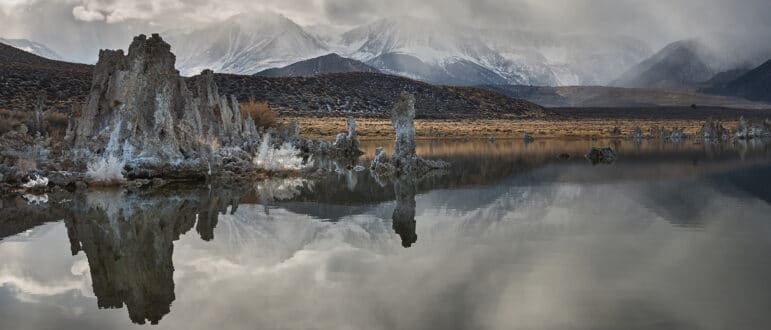 Gray clouds hang over the snowy mountains behind still, dark, tufa filled lake water.