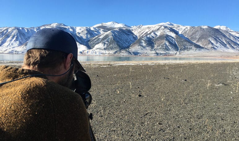 The back of a man's head as he looks at the black rock shore of Mono Lake through binoculars. Snow covered mountains are in the background.