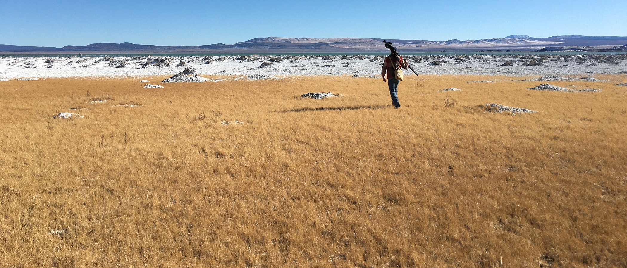 One man walks with a tripod across his back through a golden field of grass towards snow covered rocks on the shore of Mono Lake.