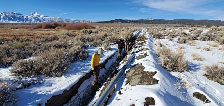 Three men are standing and walking in a long trench which stretched behind them through a snowy field of sage brush. A gray pipe sits nearby.