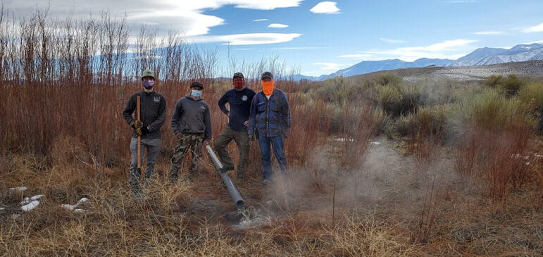 Four men in masks pose in the grasses near the pond with water flowing from a gray pipe on the ground.